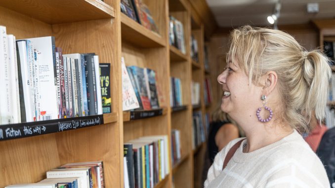 A blonde woman wearing a white top smiling at a bookshelf in a charity shop.