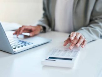 A person wearing a grey cardigan sat at a desk in front of a laptop typing on a calculator.