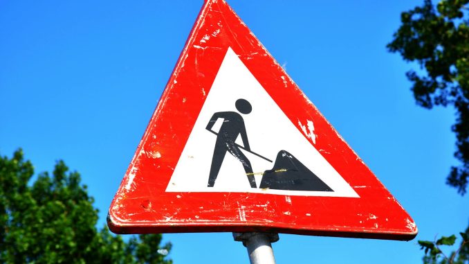 A red and white roadwork warning sign with a silhouette figure, set against a clear blue sky.