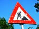 A red and white roadwork warning sign with a silhouette figure, set against a clear blue sky.