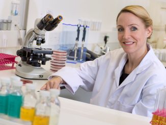 Dr Emma Saunders wearing a white lab coat, sat at a desk in a lab next to a microscope.