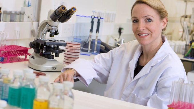 Dr Emma Saunders wearing a white lab coat, sat at a desk in a lab next to a microscope.