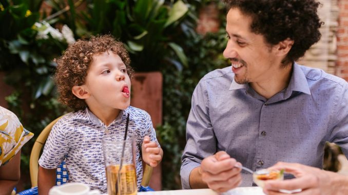Father and son share a joyful moment while dining outdoors, capturing playful expressions.